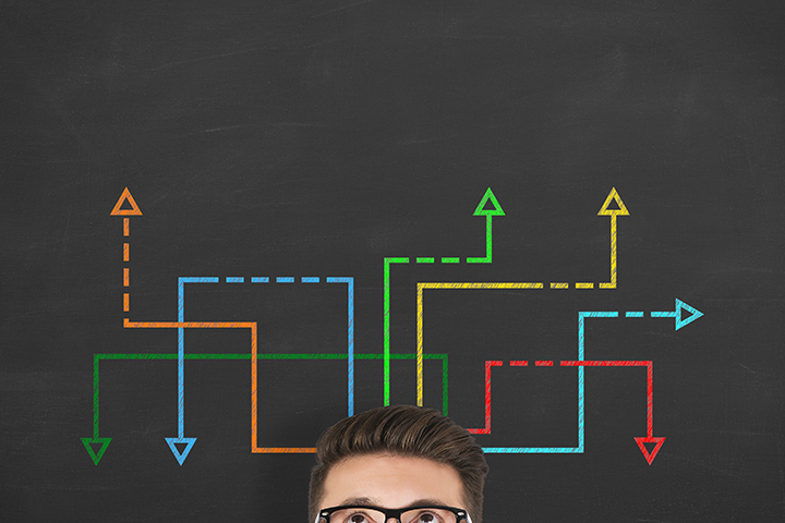 Confused, young businessman looking at many twisted arrows on the blackboard background