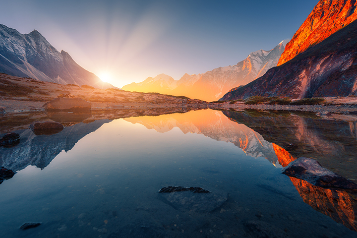 Beau paysage avec de hautes montagnes avec des pics illuminés, des pierres dans le lac de montagne, la réflexion, le ciel bleu et la lumière du soleil jaune au lever du soleil