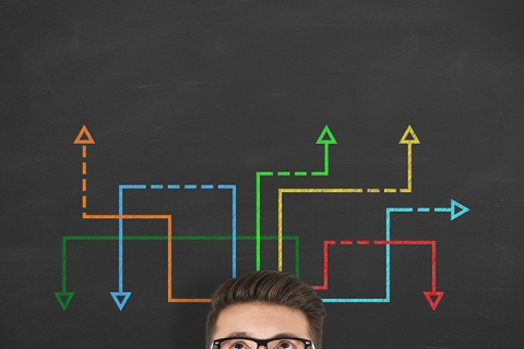 Confused, young businessman looking at many twisted arrows on the blackboard background