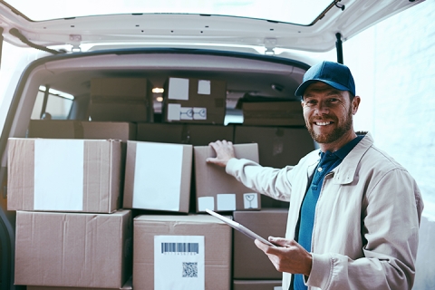 Portrait of a cheerful young delivery man standing next to a van full of boxes while holding a digital tablet