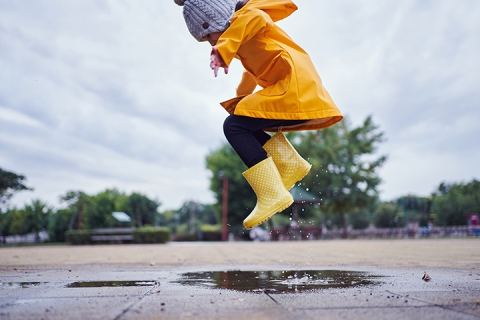 Cute and playful female child jumping in a puddle of water on the street wearing yellow rubber boots and a raincoat