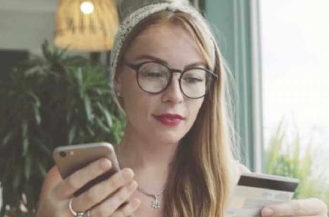 a portrait of a blond lady wearing glasses, a headband and a red lipstick holding a phone in her right hand whilst looking down at a debit card which is in her left hand