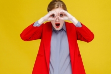 Red head woman in red suit looking in binoculars far far away. Studio shot, isolated on yellow background 
