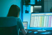 Woman in dark room looking at spreadsheets on computer screens