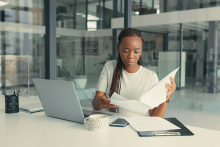 Businesswoman working at desk on laptop viewing paper documents